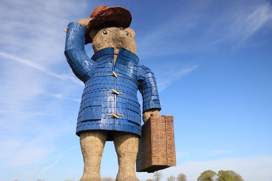 A large, stylized statue of a person with exaggerated proportions stands outdoors against a clear blue sky with a few wispy clouds. The figure has a round head with simple facial features, including small eyes, and a thick, brown hairstyle. It is dressed in a blue, textured coat that covers the torso and arms, with decorative yellow clasps along the front. The statue's legs are made from a rough, stone-like material with a beige, weathered finish, and the figure is holding a rectangular, woven brown basket or suitcase in its right hand. The scene suggests an outdoor setting, possibly near a park or open space, with distant trees visible on the horizon. The overall appearance combines artistic stylization with expressive forms, and the figure’s pose, with one hand saluting or shielding its eyes, adds a sense of character to the sculpture.