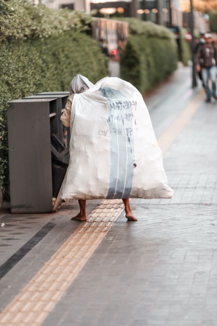 A person standing on a paved sidewalk beside a row of suitable waste bins is carrying a large, heavy-duty white sack made of thick, woven plastic material, which is partially transparent with visible blue vertical stripes and some printed text. The individual’s legs, including bare feet, are visible beneath the sack, indicating they are walking with the load. The scene is set in an urban outdoor environment, with a neatly trimmed hedge and residential or commercial buildings in the background. The sidewalk features tactile paving strips in a yellowish-brown color, running parallel along its length, which indicate a designated area for safety and accessibility. To the right, a person dressed in casual clothing is walking away or towards the scene, contributing to the sense of active pedestrian movement. The overall lighting suggests daylight, possibly in the late afternoon, with natural light illuminating the scene evenly. The setting implies a collection of domestic waste, demonstrating independent or private disposal alongside waste removal services, as seen in the context of rubbish clearance or on-site waste handling facilitated by Waste Removal Paddington.