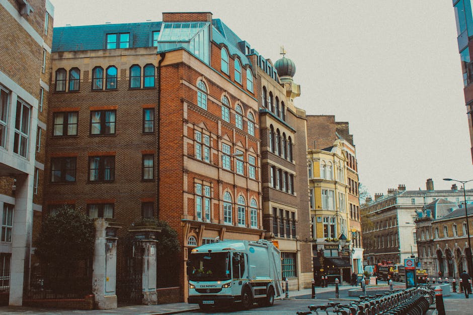 The image depicts a city street lined with multi-story buildings exhibiting a mix of architectural styles, including red brick, beige stone, and ornate detailing. In the foreground, a light blue waste collection truck with a compact cab and a rear loading area is parked on the pavement, facing towards the viewer. The truck appears clean and modern, with visible safety and operational features, and is positioned near a gated entrance that features stone pillars and black wrought iron fencing, partially obscured by some shrubbery. To the right, several bicycle racks are installed along the sidewalk, unoccupied, with a few bollards and street furniture nearby. The background shows a variety of buildings, some with large sash windows and decorative cornices, and a distinctive green-domed tower rising intermittently among the rooftops. Overhead, the sky is overcast, casting diffuse natural light over the scene. This setting suggests a private waste collection service operating in an urban area, with the vehicle parked on the street near residential or commercial buildings, exemplifying independent or alternative waste handling solutions in the city environment, aligned with rubbish removal services focused on city centre or urban locations.
