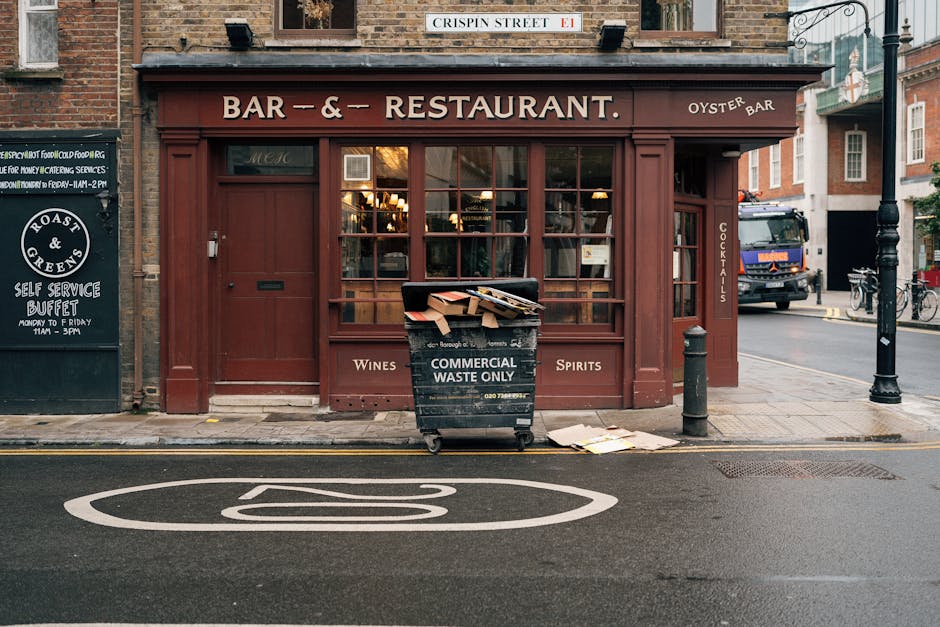 In front of a restaurant with a brown wooden façade and large glass windows, a wheeled black commercial waste bin labeled 'Commercial Waste Only' is filled with flattened cardboard boxes and paper scraps, some protruding over the top. The bin is situated on a narrow sidewalk adjacent to a designated no-parking zone, marked by a white painted symbol on the asphalt road. Nearby, there are concrete bollards and an iron street lamp, with a blackboard sign on the left advertising a self-service buffet. A delivery truck with a dark blue and orange cab is parked further along the street, and bicycles are secured to bike racks to the right. The scene is illuminated by natural daylight, with a brick building visible behind the restaurant, and a street sign reading 'Crispin Street' mounted above the shop’s fascia. The setting suggests a typical urban environment where private waste collection services, such as those provided by Waste Removal Paddington, manage refuse from local businesses, supporting alternative waste handling outside municipal waste channels.
