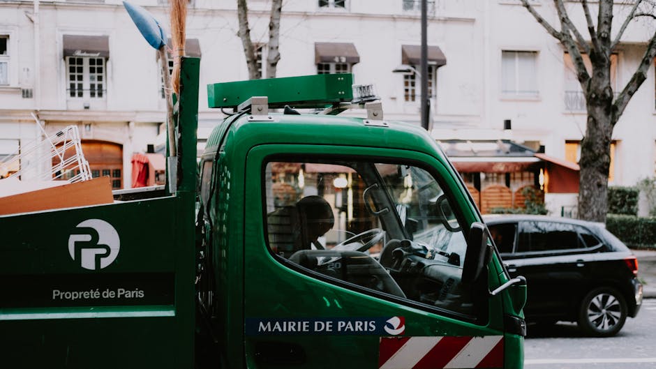 A close-up view of a green waste collection vehicle parked on a city street, with the front driver's side visible. The truck has a sleek, rounded cab with a clear windshield, revealing the steering wheel and interior details. The vehicle's side panel displays the logo of Propreté de Paris and the text 'MAIRIE DE PARIS,' indicating its municipal waste management role. Behind the truck, a black car is parked along the curb, with residential buildings featuring cream facades, multiple windows, and small balconies in the background. Leafless trees with thin branches line the street, suggesting late autumn or winter. The scene is evenly lit with natural daylight, and the overall setting reflects an urban environment engaged in waste collection, which could relate to private waste handling services like those provided by Waste Removal Paddington for commercial or residential rubbish disposal.