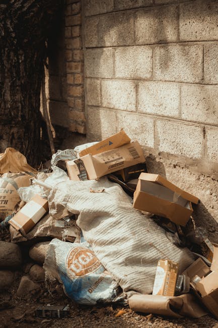 A pile of discarded cardboard boxes, some flattened and others partially intact, along with plastic bags and packaging materials, are accumulated at the base of a rough-textured brick wall and a tree trunk. The cardboard boxes are light brown with visible printed labels and barcode stickers, and some are slightly torn or crumpled. The plastic bags appear to contain additional waste and are partially covered by a coarse, woven fabric or sackcloth. The scene is outdoors, with the waste resting on uneven ground consisting of dirt, small stones, and debris, near a brick wall that provides a backdrop. The lighting suggests natural daylight, casting subtle shadows on the waste and wall, indicating an environment where private or independent rubbish disposal might be taking place. Waste removal professionals such as those from Waste Removal Paddington might respond to such waste accumulation, providing a clean-up or alternative waste management service, especially when dealing with household or construction debris outside typical council collection areas.