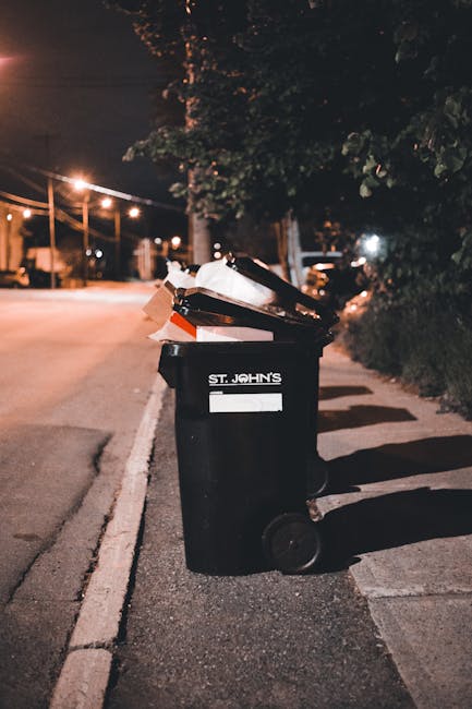 A black wheelie bin with a white label and the words 'ST. JOHN'S' printed on the front, positioned on a city pavement at night. The bin is filled with various types of rubbish, including cardboard boxes, plastic bags, and other waste items. The bin's lid is open, revealing the contents inside. It stands on a sidewalk next to a curb, with a row of similar bins extending into the background. The scene is illuminated by streetlights, casting shadows on the ground and highlighting the textured surface of the bin and surrounding pavement. Behind the bins, there is a dark, leafy hedge and a tall tree with dense foliage partially visible. In the distance, faint streetlights and parked cars can be seen along the road, emphasizing an urban environment consistent with private waste collection and rubbish removal services like those offered by Waste Removal Paddington.