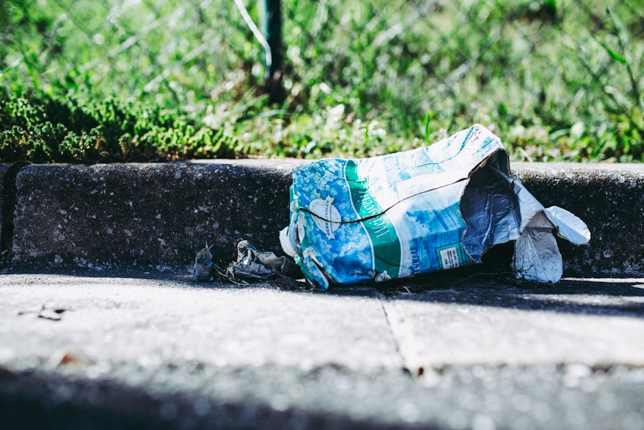 A pile of discarded cardboard boxes, some flattened and others partially intact, along with plastic bags and packaging materials, are accumulated at the base of a rough-textured brick wall and a tree trunk. The cardboard boxes are light brown with visible printed labels and barcode stickers, and some are slightly torn or crumpled. The plastic bags appear to contain additional waste and are partially covered by a coarse, woven fabric or sackcloth. The scene is outdoors, with the waste resting on uneven ground consisting of dirt, small stones, and debris, near a brick wall that provides a backdrop. The lighting suggests natural daylight, casting subtle shadows on the waste and wall, indicating an environment where private or independent rubbish disposal might be taking place. Waste removal professionals such as those from Waste Removal Paddington might respond to such waste accumulation, providing a clean-up or alternative waste management service, especially when dealing with household or construction debris outside typical council collection areas.