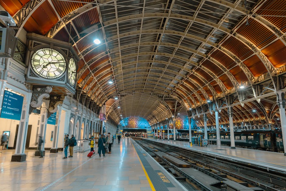 The image shows a spacious indoor train station with a high, arched metal and glass roof supported by ornate white columns. On the left side, there is a large decorative clock with Roman numerals, encased in a vintage-style frame, mounted on the wall above blue information signs. The station floor is tiled with grey and beige patterns, and a few travelers are visible, some with luggage, walking or standing on the platform. The platform is clear and extends into the distance, with train tracks running alongside. Interior lighting creates a warm ambiance, contrasting with the cooler blue-toned lights seen at the far end of the station's arched ceiling. The scene conveys a clean, well-maintained environment suitable for passenger transport, with no visible rubbish or debris. As a professional waste management service provider, Waste Removal Paddington can assist with maintaining such areas free from unwanted waste, ensuring safe and efficient travel environments. This setting highlights the importance of regular, independent waste collection to support the cleanliness and functionality of large transportation hubs.
