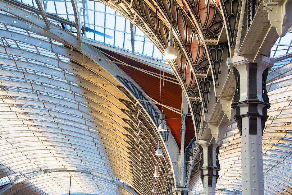 The image depicts the interior of an ornate, historic railway station with an arched, vaulted ceiling constructed of white-painted iron and glass. The ceiling features intricate metal trusses and supports, painted in a combination of cream, black, and dark brown, which contribute to the architectural detail and structural stability. Large glass panels allow natural daylight to illuminate the space, casting soft reflections on the polished surfaces below. Suspended from the ceiling are modern metallic pendant lights, providing additional artificial lighting. Visible in the foreground are parts of the supporting iron columns, with riveted joints and decorative detailing, highlighting the station’s period design. The overall atmosphere is bright and airy, showcasing the blend of historical craftsmanship and natural light. This architectural style aligns with the context of alternative, on-site waste handling and private collection solutions often utilized within large public or historic transportation hubs, exemplifying a space where artificial and natural lighting combine to illuminate operational areas associated with station maintenance or service facilities, such as waste removal or storage areas. Waste Removal Paddington occasionally services such environments for efficient rubbish management.
