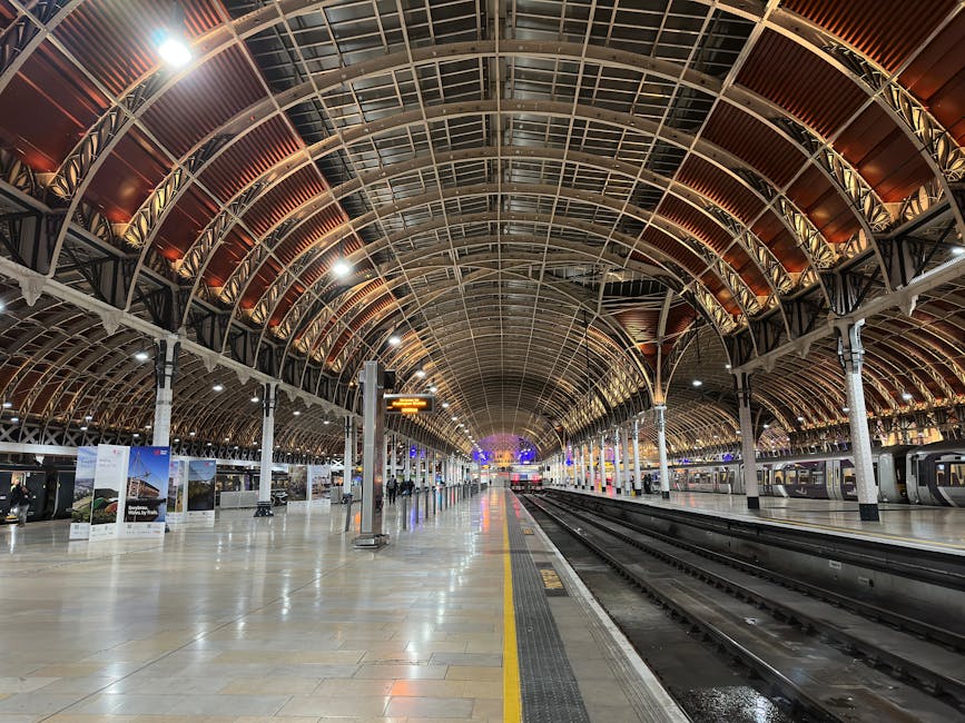 The image depicts the arching roof structure of Paddington Station, showcasing a large, semi-circular glass and metal canopy with intricate white ironwork detailing. The transparent glass panels reflect a partly cloudy blue sky with fluffy white clouds, illuminating the station's entrance area. Prominently centered on the white section of the arch is the station name, 'Paddington Station,' written in clear blue lettering. At the very top of the arch is an ornate coat of arms featuring red, white, and blue elements, adding a historic or official decorative touch. The surrounding environment hints at a busy railway station façade, with the partial edge of an adjacent building visible on the left side, constructed from grey stone or brick. The overall atmosphere is bright and well-lit, emphasizing the architectural details and the open, inviting structure of the station entrance, which is associated with transportation hubs where various forms of public and private transportation, including independent waste collection services like those of Waste Removal Paddington, typically operate in the vicinity to manage waste from travelers and station activities.