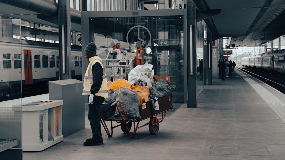 A man dressed in a dark jacket, beanie hat, and white gloves stands on a modern train station platform next to a large, red, metal trolley loaded with black, grey, yellow, and white trash bags, some of which are tied or partially open, revealing waste inside. The trolley is positioned close to a grey waste container or bin, and behind it, a glass-walled elevator or kiosk structure features mechanical equipment visible through the transparent panels. In the background, there are parked train carriages with closed doors on an adjacent track, and a few other figures are visible further along the platform. The platform surface is composed of large, smooth, grey tiles, and the scene is illuminated by natural daylight, creating a neutral, professional atmosphere suited to the context of waste or rubbish handling within a transportation hub. The image subtly reflects the theme of independent waste collection or on-site rubbish management, relevant to private waste removal services such as those offered by Waste Removal Paddington.