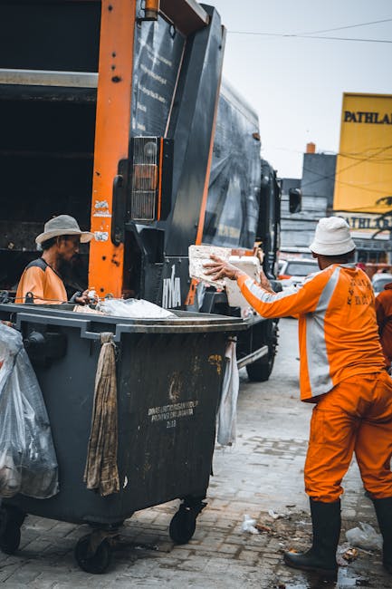 An outdoor scene showing two waste management workers in high-visibility orange uniforms and white helmets, engaged in rubbish collection. One worker stands on the right side, holding a large piece of discarded material, possibly packaging or a flat sheet, about to place it into a black wheelie bin positioned in front of a large refuse truck. The other worker on the left quietly observes, with a gloved hand resting on the bin, which has a wooden handle or guard attached to its side. The refuse truck behind them is primarily dark with orange accents, equipped with a hydraulic lift system for waste collection, and part of its loading mechanism is visible on the left. The scene takes place on a paved urban street with a yellow advertising sign partially visible in the background, alongside buildings and parked vehicles, indicative of an active area for private waste collection. The natural lighting suggests daytime, with shadows cast on the ground, emphasizing the urban environment's functional aesthetic. Waste Removal Paddington provides services such as on-site rubbish clearance, evident from the scene of active waste handling and collection in a busy vicinity. This image exemplifies an independent collection process for bulky or general waste, highlighting the practical aspect of private disposal methods aligned with non-local authority rubbish removal. The overall composition focuses on the interaction between workers and waste equipment within a typical city street setting, supporting the topic of alternative waste handling. The level of detail includes the textural contrast of the plastic waste, the reflective safety gear, and the utilitarian design of the collection vehicles, reinforcing the professional approach to rubbish removal. 