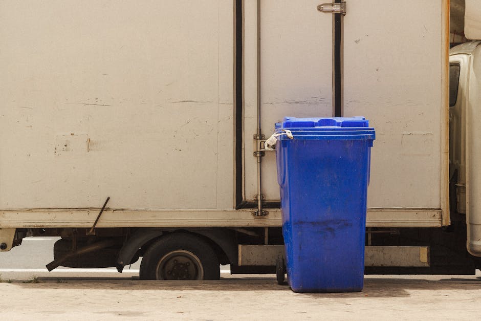 An outdoor scene showing two waste management workers in high-visibility orange uniforms and white helmets, engaged in rubbish collection. One worker stands on the right side, holding a large piece of discarded material, possibly packaging or a flat sheet, about to place it into a black wheelie bin positioned in front of a large refuse truck. The other worker on the left quietly observes, with a gloved hand resting on the bin, which has a wooden handle or guard attached to its side. The refuse truck behind them is primarily dark with orange accents, equipped with a hydraulic lift system for waste collection, and part of its loading mechanism is visible on the left. The scene takes place on a paved urban street with a yellow advertising sign partially visible in the background, alongside buildings and parked vehicles, indicative of an active area for private waste collection. The natural lighting suggests daytime, with shadows cast on the ground, emphasizing the urban environment's functional aesthetic. Waste Removal Paddington provides services such as on-site rubbish clearance, evident from the scene of active waste handling and collection in a busy vicinity. This image exemplifies an independent collection process for bulky or general waste, highlighting the practical aspect of private disposal methods aligned with non-local authority rubbish removal. The overall composition focuses on the interaction between workers and waste equipment within a typical city street setting, supporting the topic of alternative waste handling. The level of detail includes the textural contrast of the plastic waste, the reflective safety gear, and the utilitarian design of the collection vehicles, reinforcing the professional approach to rubbish removal. 