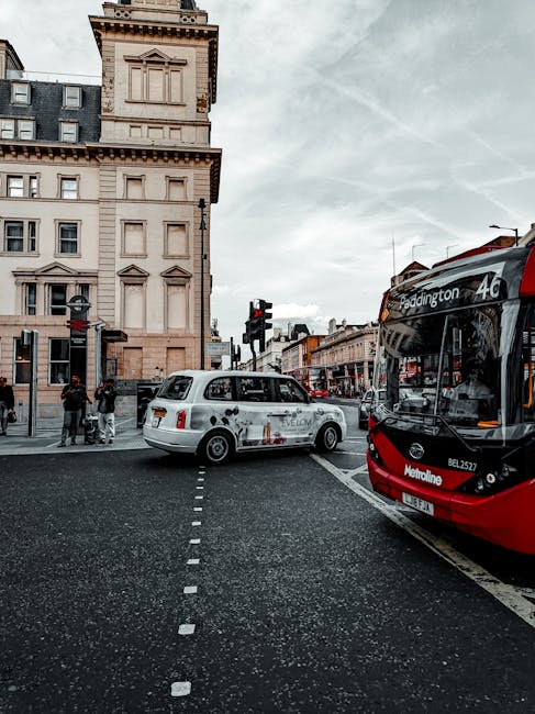 A white taxi with a black roof and advertising decals on the sides is stopped partially across a pedestrian crossing on a city street. Behind the taxi, a group of four pedestrians, including two adults and two children, are waiting to cross near a traffic light. To the right, a red and black modern double-decker bus displaying route information for Paddington is parked at a bus stop. In the background, there is a tall, beige, multi-story building with decorative stonework, rectangular windows, and a prominent tower-like section with architectural detailing. The sky overhead is overcast with light gray clouds, creating diffuse lighting that reduces shadows. The street scene, captured from eye level, reflects a typical urban landscape where public transport and private vehicles are common, and it is situated in an area managed by Westminster Council. This setting subtly relates to the broader context of city waste management, including rubbish removal practices observed on urban streets managed under local council regulations, or those related to private waste services such as Waste Removal Paddington.
