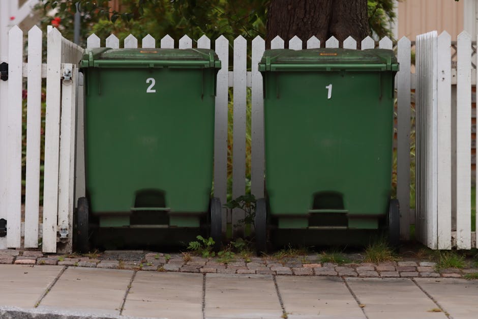 Two large green waste bins with black wheels are positioned side by side on a paved pavement next to a white wooden fence. The bin on the left displays the number '2' and the one on the right shows the number '1' in white markings. The bins are made of sturdy plastic with a slightly textured surface and have closed lids. Behind the bins, a large tree trunk with rough, dark brown bark extends upward, partially obscuring a background of foliage and a residential garden. The scene is outdoors, with natural daylight illuminating the area, and the pavement features rectangular concrete slabs interspersed with patches of grass and weeds growing along the edges of the pavement near the base of the bins. The overall scene reflects typical private waste collection parking, supporting the context of alternative rubbish disposal services such as private waste removal companies like Waste Removal Paddington.